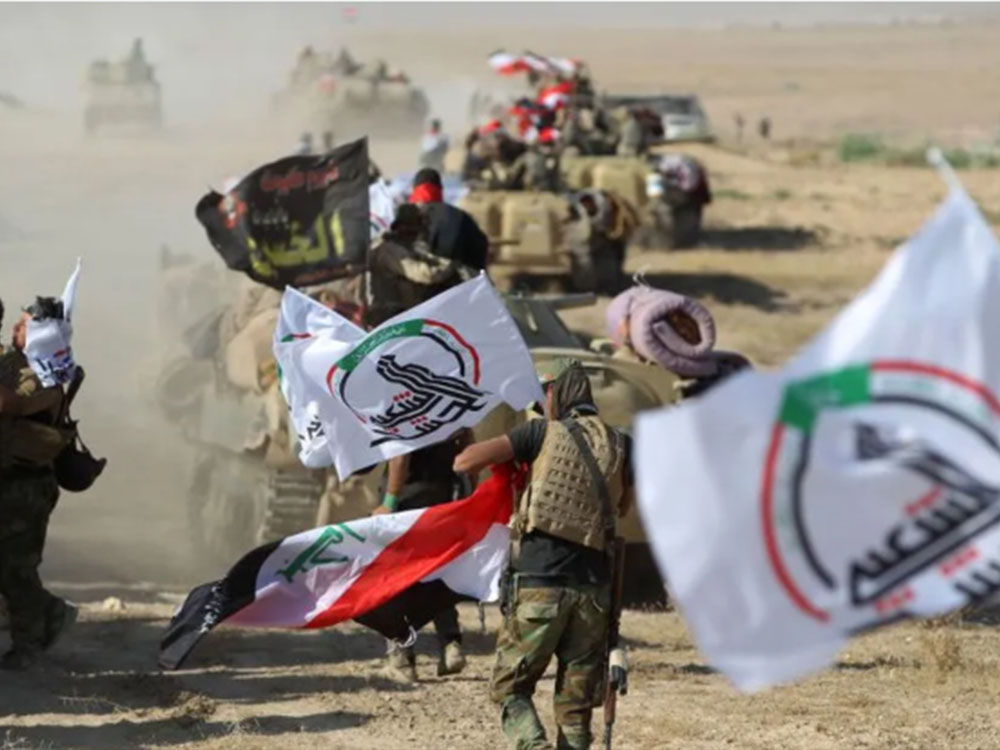 عناصر من هيئة الحشد الشعبي يشاركون في تحرك ميداني ضمن رتل عسكري في منطقة صحراوية، وترفرف أعلام العراق ورايات الهيئة على طول مسار الآليات العسكرية.