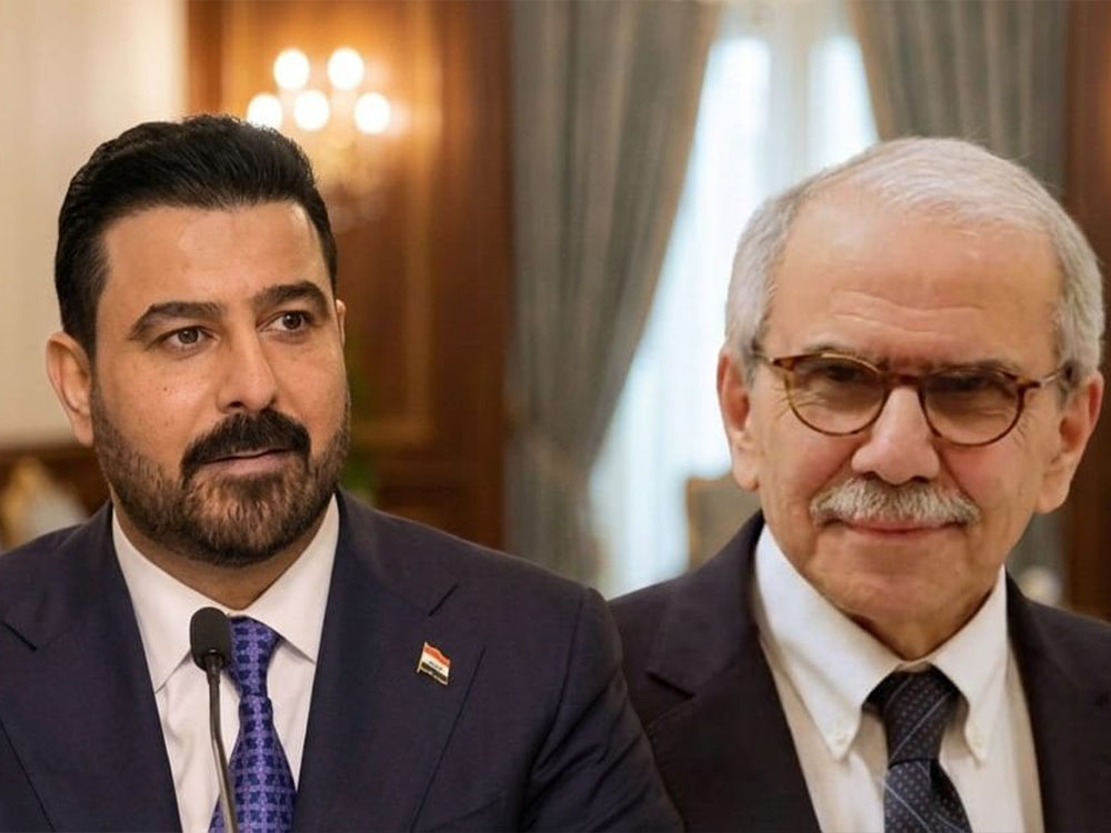 رجلان يرتديان بدلتين رسميتين يظهران داخل قاعة اجتماعات، أحدهما يتحدث أمام ميكروفون والآخر يجلس إلى جواره مبتسماً.