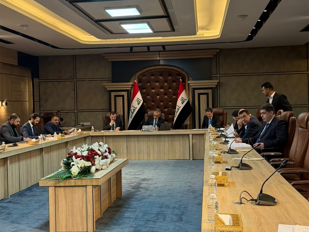قاعة اجتماعات رسمية تضم اللجنة المالية النيابية يجلسون حول طاولة مستطيلة، تتوسطها باقة زهور، فيما تظهر الأعلام العراقية خلف رئيس الجلسة.