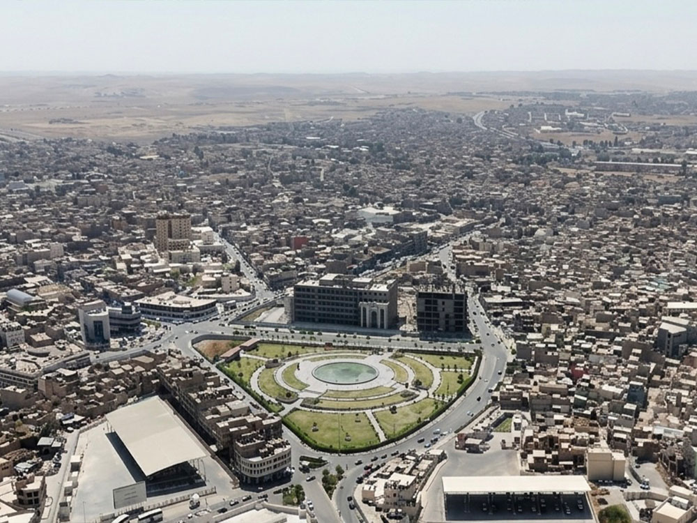 صورة جوية لمدينة الموصل تُظهر ساحة الاحتفالات والمباني الحكومية المحيطة بها، مع شبكة الطرق والأحياء السكنية الممتدة في مختلف أنحاء المدينة.