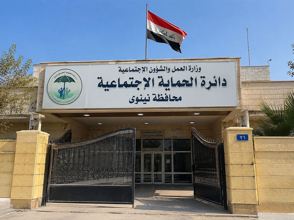 واجهة مبنى دائرة الحماية الاجتماعية في محافظة نينوى، يتوسطها المدخل الرئيسي ويرتفع فوقها العلم العراقي.