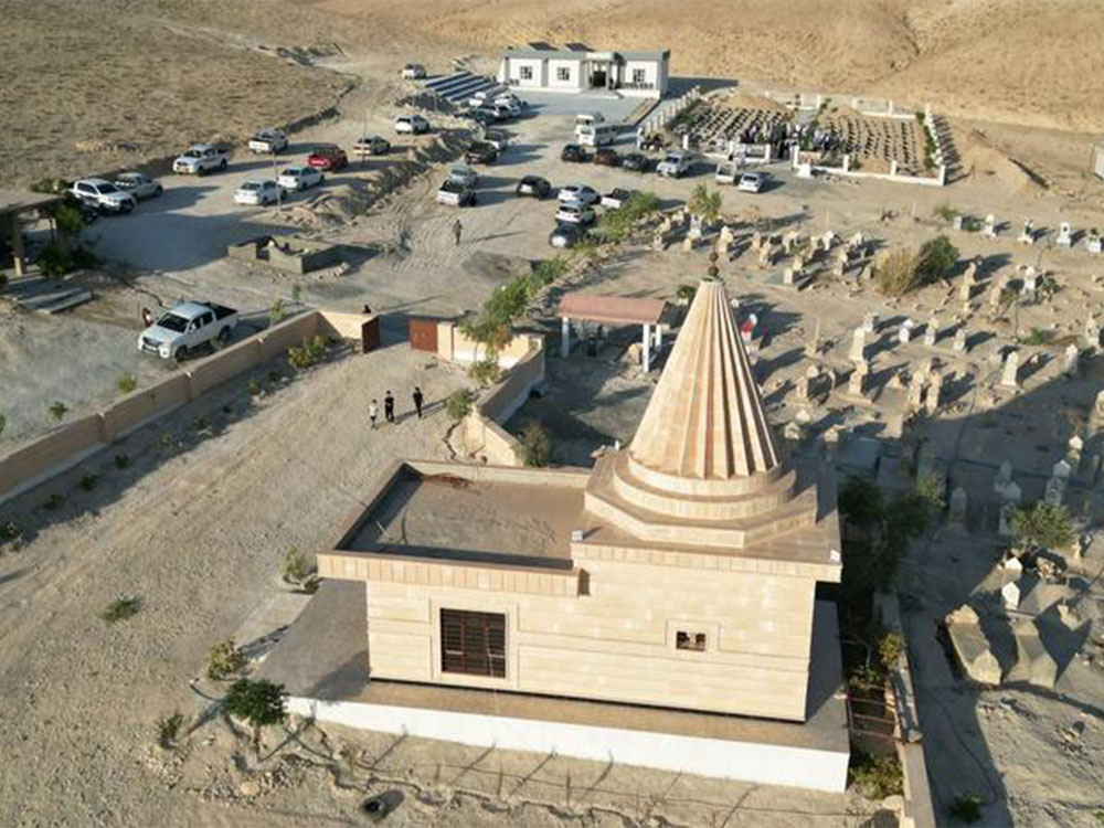 لقطة جوية لمزار ديني إيزيدي في منطقة سنجار بمحافظة نينوى، يظهر المبنى ذو القبة المخروطية المميزة محاطًا بمقبرة وساحة استقبال ومركبات الزائرين، وسط طبيعة جبلية جافة.