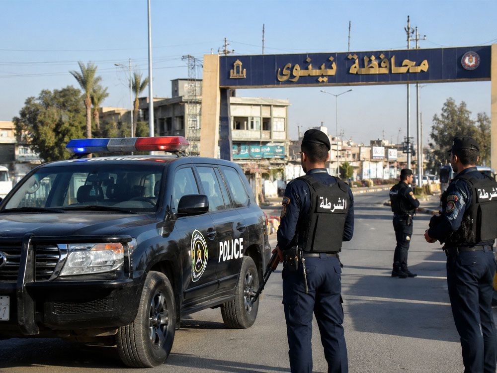 عناصر من الشرطة العراقية يقفون إلى جانب مركبة أمنية عند أحد مداخل محافظة نينوى في مدينة الموصل، ضمن مشهد يرمز إلى الإجراءات الأمنية وتنفيذ الواجبات الرسمية.