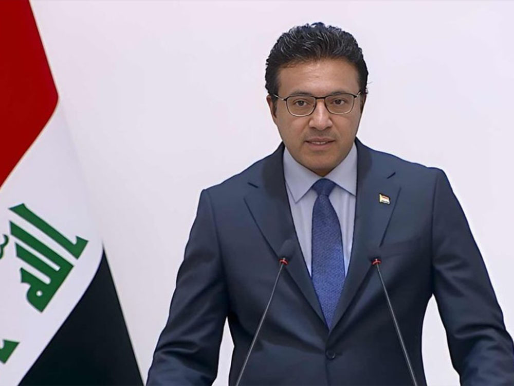 المتحدث باسم الحكومة العراقية حيدر العبودي يقف أمام منصة خلال مؤتمر رسمي، وخلفه العلم العراقي.