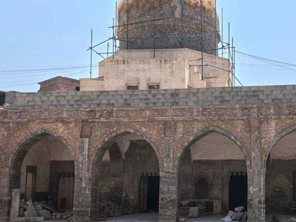 صورة لجامع القطانين التاريخي، تُظهر رواقاً أثرياً بأقواس إسلامية مدببة وبناءً حجرياً قديماً يعلوه جدار حديث من البلوك الخرساني، وتظهر في الخلفية قبة دائرية قيد الترميم محاطة بسقالات معدنية تحت سماء صافية