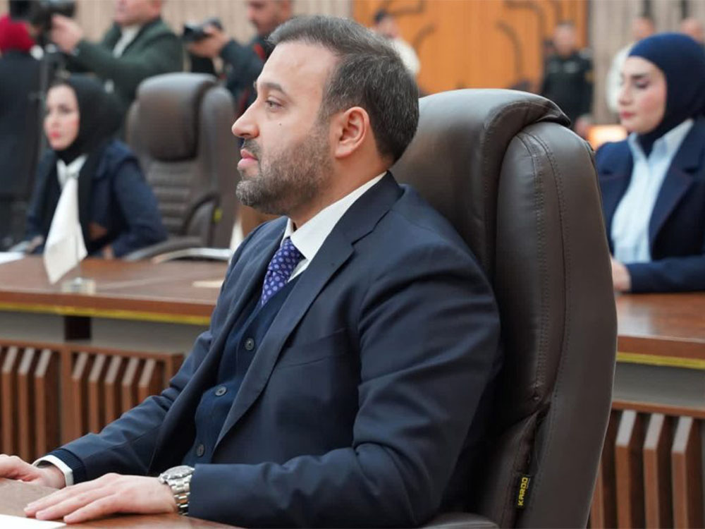 لقطة جانبية لرجل يرتدي بدلة كحلية جالسًا على كرسي مكتب جلدي كبير في قاعة مؤتمرات مزدحمة، ينظر إلى اليسار بعيدًا عن الكاميرا.