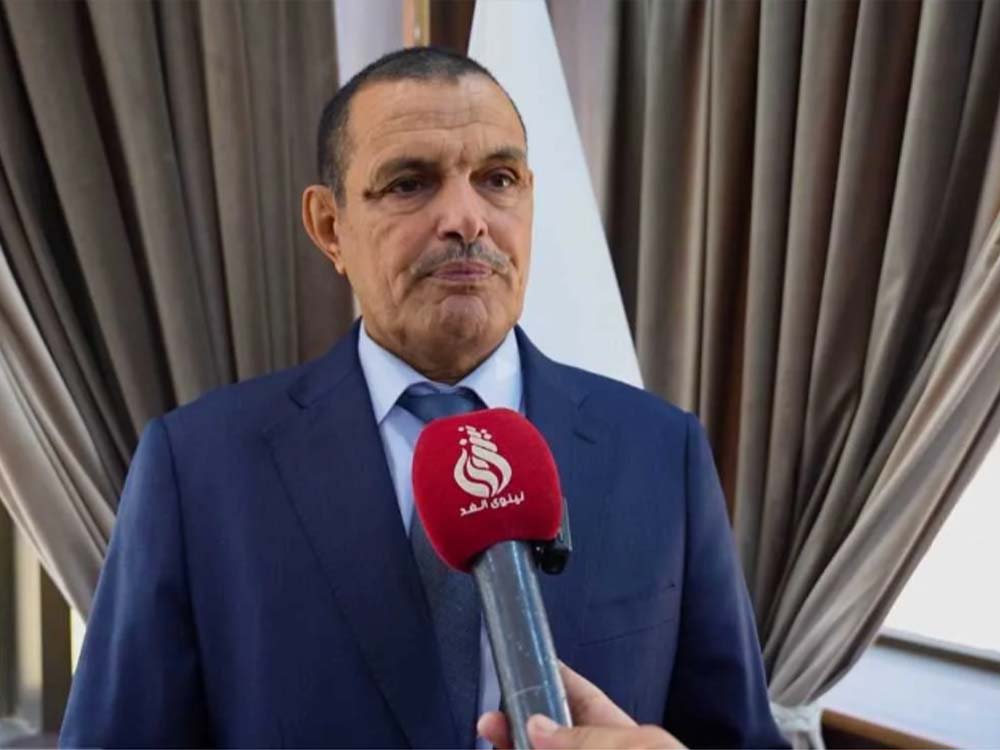 عضو مجلس محافظة نينوى محمد هريس يتحدث خلال مقابلة صحفية، مرتدياً بدلة رسمية زرقاء، بينما يحمل مراسل ميكروفوناً أحمر أمامه داخل إحدى القاعات.