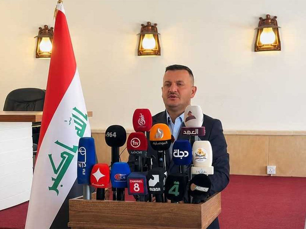 مدير عام آثار وتراث نينوى يتحدث خلال مؤتمر صحفي، محاطًا بعدد من ميكروفونات وسائل الإعلام، وبجانبه العلم العراقي.