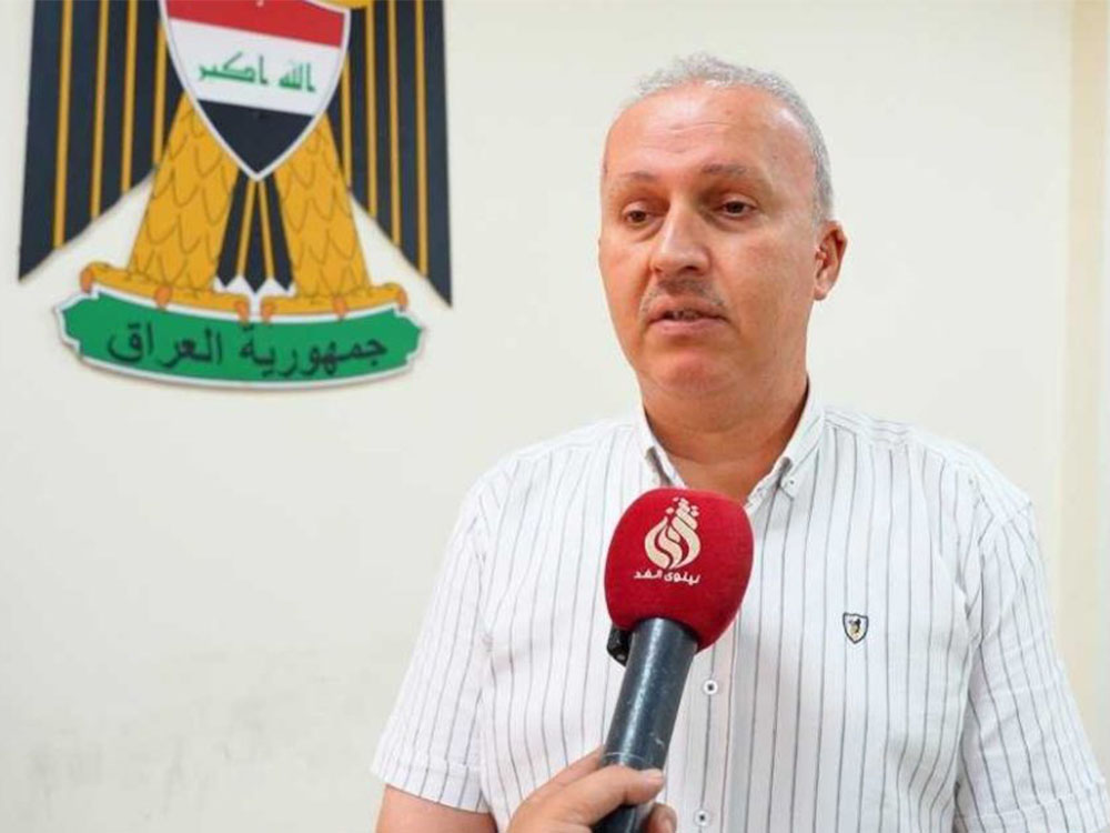 "صورة لشخص مسؤول يتحدث أمام ميكروفون أحمر يحمل شعار قناة 'نينوى الغد'، مرتدياً قميصاً أبيض مخططاً بخطوط رفيعة، وعلى الجدار خلفه يظهر شعار جمهورية العراق (النسر الجمهوري)."