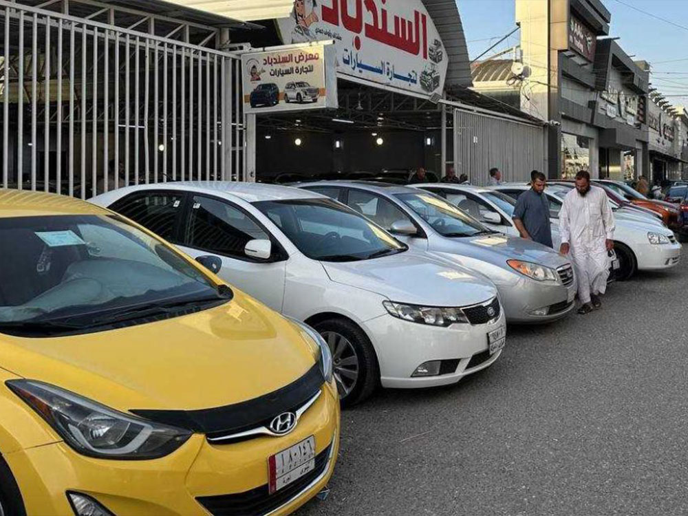 صف من السيارات الجديدة والمستعملة معروضة أمام أحد معارض السيارات في مدينة الموصل، فيما يتجول عدد من المواطنين بين المركبات.