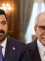 رجلان يرتديان بدلتين رسميتين يظهران داخل قاعة اجتماعات، أحدهما يتحدث أمام ميكروفون والآخر يجلس إلى جواره مبتسماً.