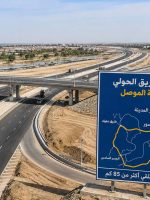 مشهد جوي لطريق سريع حول مدينة الموصل يضم جسوراً وتقاطعات متعددة، مع حركة شاحنات ومركبات ولوحة توضيحية لمسار الطريق الحولي.