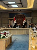 قاعة اجتماعات رسمية تضم اللجنة المالية النيابية يجلسون حول طاولة مستطيلة، تتوسطها باقة زهور، فيما تظهر الأعلام العراقية خلف رئيس الجلسة.