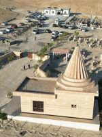 لقطة جوية لمزار ديني إيزيدي في منطقة سنجار بمحافظة نينوى، يظهر المبنى ذو القبة المخروطية المميزة محاطًا بمقبرة وساحة استقبال ومركبات الزائرين، وسط طبيعة جبلية جافة.