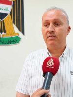 "صورة لشخص مسؤول يتحدث أمام ميكروفون أحمر يحمل شعار قناة 'نينوى الغد'، مرتدياً قميصاً أبيض مخططاً بخطوط رفيعة، وعلى الجدار خلفه يظهر شعار جمهورية العراق (النسر الجمهوري)."