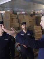 مسؤول يتحدث ويشير بيده إلى ضابط شرطة بحضر ضابط آخر، داخل مستودع مليء بالصناديق الكرتونية المكدسة خلال جولة تفتيشية.